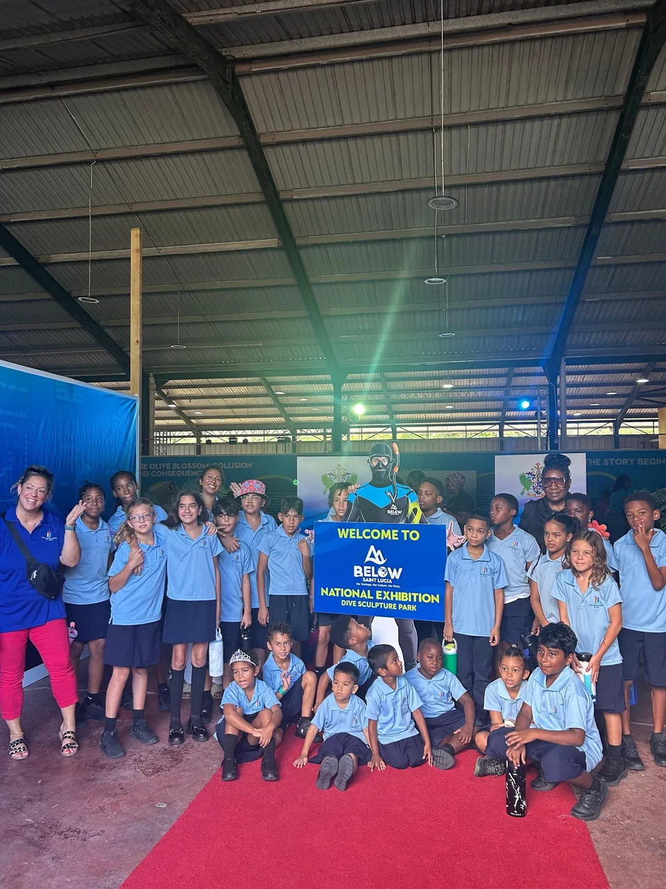 Students and a teacher pose on a red carpet behind a 'Welcome to Below National Exhibition' sign