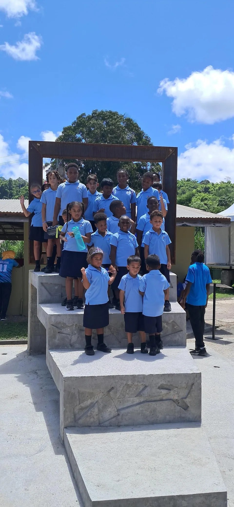 Younger students stand on stone steps under a wooden frame outdoors beneath a blue sky