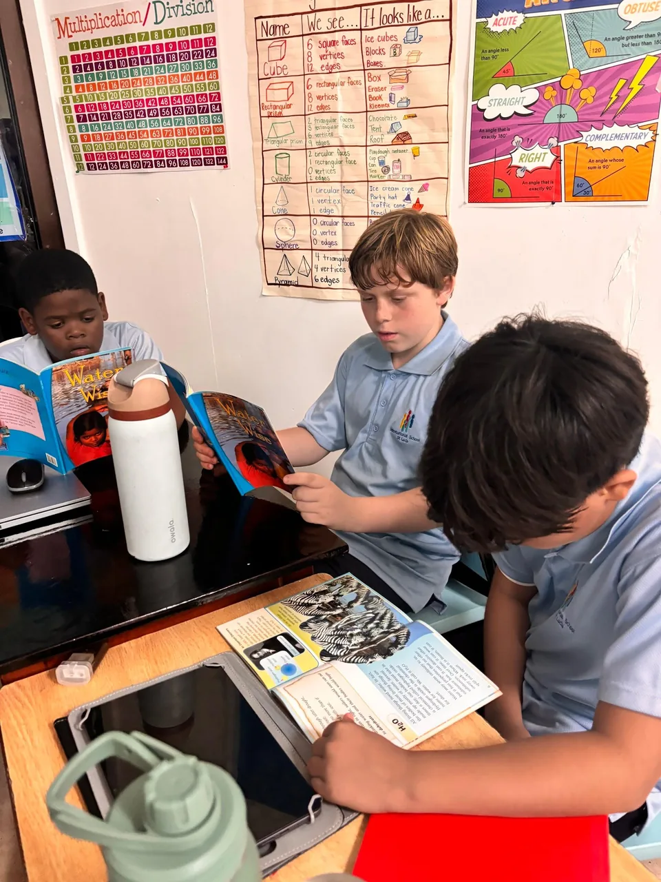 Three boys read books at a desk beneath maths posters