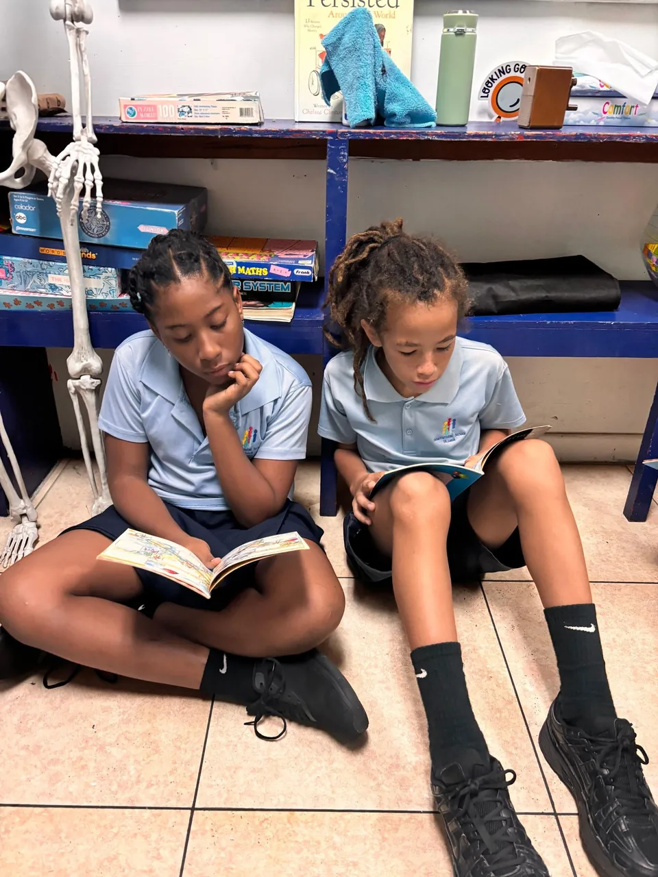 Two students sit on the floor reading books next to a skeleton model
