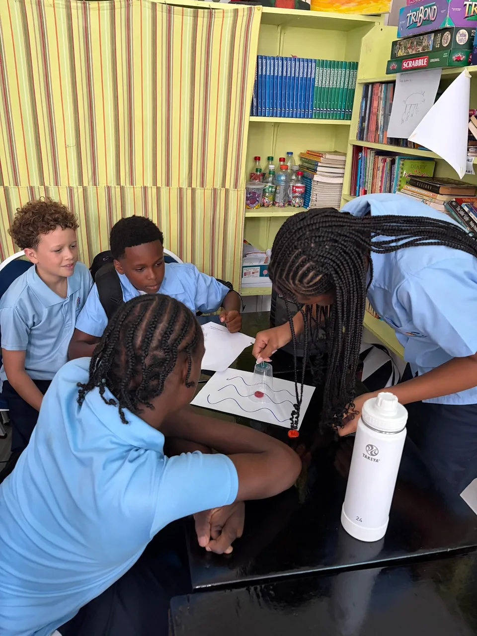 A student guides a small cup across a wavy-line sheet as classmates watch