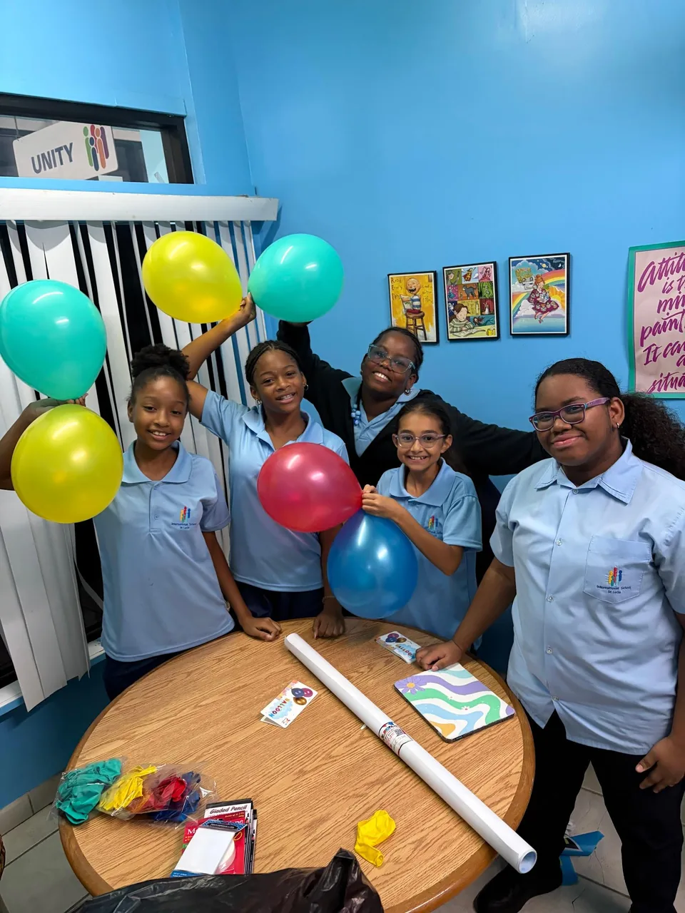 Smiling students hold up colourful balloons around a classroom table