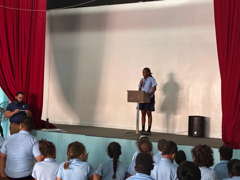 A student speaks at a lectern on the hall stage as seated students and a teacher listen