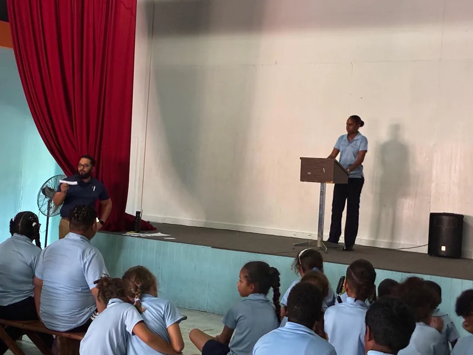A candidate stands at the lectern while a teacher with notes and seated students look on