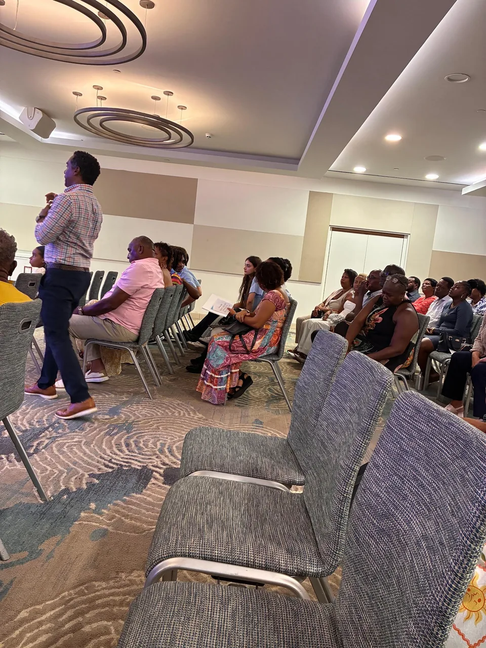 Audience seated in a hotel conference room while a man stands and speaks during the symposium