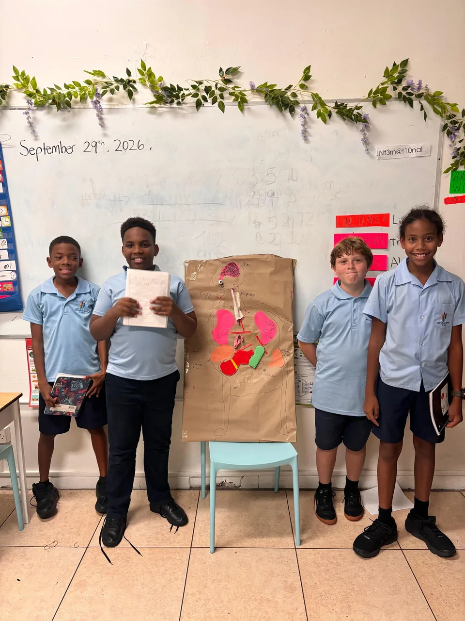 Four students present their brown-paper body poster in front of the classroom whiteboard
